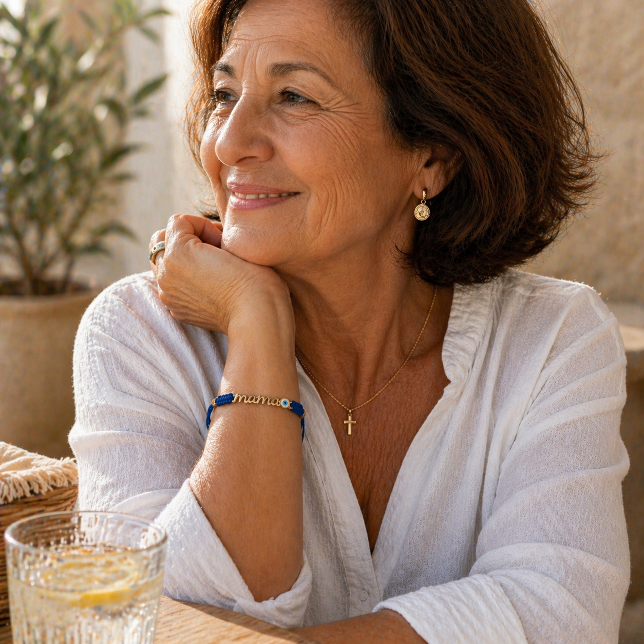 Woman in a white shirt sitting outdoors with a glass of water, wearing a mama bracelet with Evil Eye