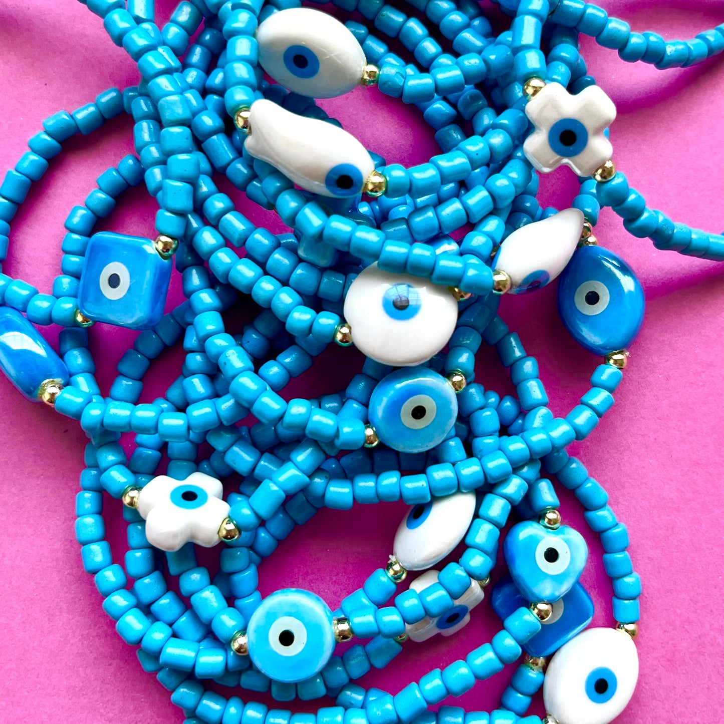 Mykonos mati stackable bracelets with ceramic evil eye charms. Blue beaded necklace with white eye-shaped beads on a pink background. 
