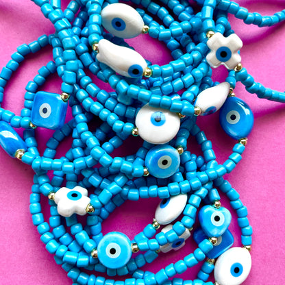 Mykonos mati stackable bracelets with ceramic evil eye charms. Blue beaded necklace with white eye-shaped beads on a pink background. 